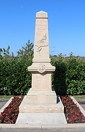 Un monument aux morts blanc en forme d'obélisque entouré par des fleurs rouges à sa base.