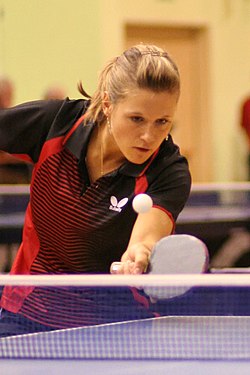 Atleta loira veste camisa vermelha e preta e, segurando uma pequena raquete, olha atentamente para a bola, que passa sobre a rede colocada no meio de uma mesa azul.