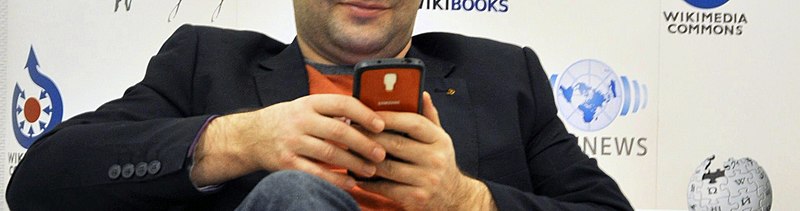 A person holds a mobile phone; in the background are Wikimedia project logos