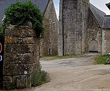 Photographie en couleurs d'un pilier de clôture couvert de végétations, d'anciens bâtiments en pierre visibles en arrière-plan.