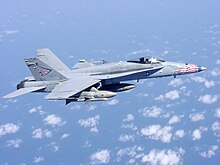 Overall-gray jet fighter, with red, blue and white-tipped nose, overflying sea and scattered white clouds down below. The aircraft is carrying streamlined external fuel tanks and missiles under its wings, and is heading right.