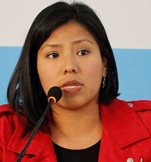 Das Foto zeigt Indira Huilca, die nach vorne gerichtet in ein Mikrofon spricht. Sie hat mittellange schwarze Haare und trägt einen roten Mantel über ein schwarzes Oberteil. Der Hintergrund ist blau-grau.