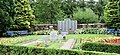 Photo d'un montrant un jardin avec des buissons bien taillés et des allées de fleurs de chaque côté. Au milieu se trouve des pierres commémoratives et dans le fond un monument en trois parties avec l'inscription du nom des victimes.