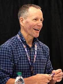 A three-quarters bust portrait photo shows a middle-aged white man looking to the photographer's right; he is wearing a blue-plaid collared shirt and a lanyard.