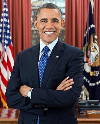 Barack Obama standing in front of a wooden writing desk and two flagpoles