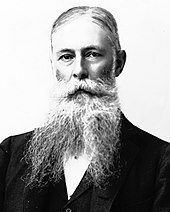 portrait shot of a man of about sixty, with hair combed across his head and with a beard divided in two. He wears a suit coat but his neckwear is obscured by the beard.