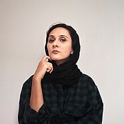 Fatemeh Behboudi ist mit einem dunklem Kopftuch zu sehen, aus dem ihr dunkles Haar erkennbar ist. Sie schaut ernst blickend in die Kamera und hat die rechte Hand mit gespreiztem Zeigefinger am Kinn. Der Hintergrund ist hell.