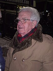 Пожилой белый мужчина с седыми волосами в очках в теплом пальто и шарфе, изображенный на улице ночью.