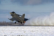 Three-quarter hind view of grey jet aircraft taking off in snowy environment. Its engine is kicking up snow on the ground