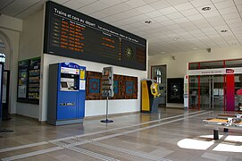 Hall d'accueil vide avec le panneau d'indication des trains sur le mur et les automates en-dessous. L'accès aux guichets est au fond.