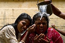Duas mulheres de cor parda e cabelos negros. Na da direita, de roupa vermelha, está sendo despejado um balde de água sobre sua cabeça, por isso exibe uma expressão de espanto. A mulher da direita, de vestes brancas, sorri.
