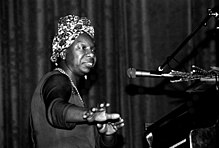 Head and torso of black woman at a piano in front of a microphone wearing a flowered kerchief in her hair