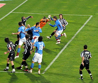 Joueurs de football dans la surface de réparation se disputant un ballon aérien pendant que le gardien s'étend dans les airs pour tenter aussi d'attraper la balle