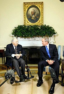 An old man in a wheel chair is talking to a middle-aged man sitting to the right. In the background, above their heads are a plant decoration and a portrait of some historical person.