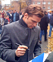 Мужчина в темно-сером шерстяном пальто на улице дает фанатке автограф.