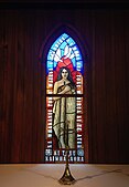 Lady Chapel stained glass window, commissioned in 2004. A wahine (woman) draped in a korowai with pounamu cross necklace holding Te Paipera Tapu (the Māori Bible) framed by Luke 1:46-47. Below her are crests of Te Pīhopatanga o Te Tai Tokerau. Above her are doves.