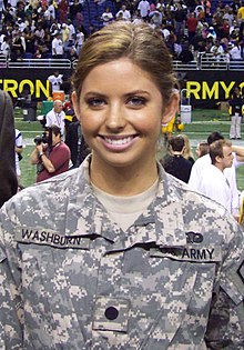 A bust photo of a white woman with brown hair; she is wearing a grey, camouflage US Army uniform and smiling into the camera