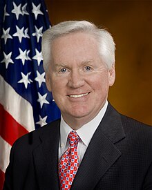 A photographic portrait of an older man from the shoulders up, wearing a suit, with a necktie, in front of an American flag