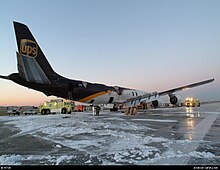 Rear-quarter view of a large aircraft with visible fire damage parked on an area of airport tarmac covered in patches of firefighting foam.