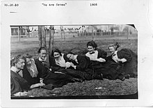 Seven women dressed in 1900s attire, laying in the grass on campus