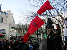 Три красных флага с логотипами IWW несут над толпой людей.