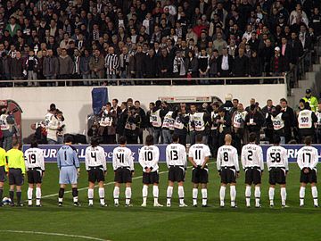 Joueurs de football en blanc alignés sur le terrain face à une des tribunes du stade