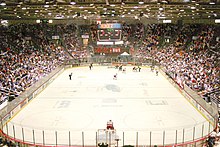 The view of an ice hockey rink from behind and above one goal. Most players, dressed in either red and white or blue and black, are in the far right corner of the rink. Above center ice an electronic scoreboard reads "9:09" in green; under that, red numbers read from left to right "0", "1", and "0". The "1" is smaller than the two "0"s. On the visible end of the rink, a red sign reads in the black and white text "Pizza Hut".