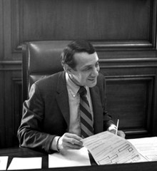 黑白照片，照片中一名男子身着正装，坐在桌旁翻阅文件