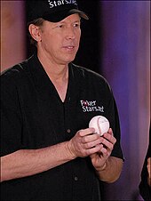 A tanned male baseball player in his fifties wearing a black cap and shirt, holding a baseball.