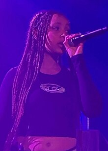 In hazy blue lighting, PinkPantheress performs with a mic in her hand.