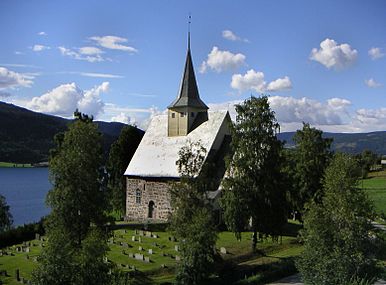 Malý kamenný kostelík s malou věží na dřevěné zvonici sedí na zeleném hřbitově s výhledem na jezero a hory.