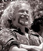 Black and white photo of a man in his fifties, looking to the right, smiling and folding his arms at his chest. He has curly, light-colored hair and a goatee, and is wearing a flower-patterned shirt with rolled-up sleeves.