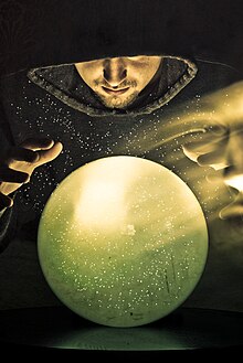 Colour photograph of a hooded wizard gazing into a green crystal ball with the ghost image of his face floating to the right.