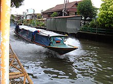 A boat, about mười mét (33 foot) long, travelling along a canal, the dark water breaking up in foam as it passes