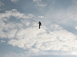 A man walks across a tightrope, with only a clouded sky in the background
