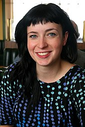 Photographie en buste d’une jeune femme souriante aux cheveux noirs qui regarde l’objectif.