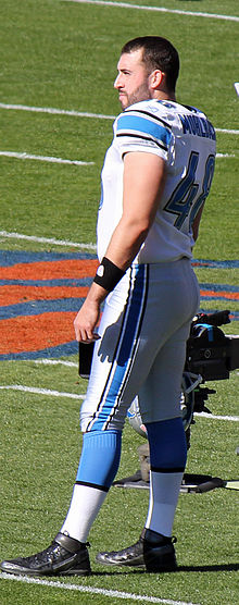 Side view of Don Muhlback in his full Detroit Lions uniform while holding his helmet in his hand.