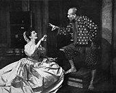 woman kneeling in front of a standing man; the two are conversing and each is gesturing with one hand as if ringing a small bell