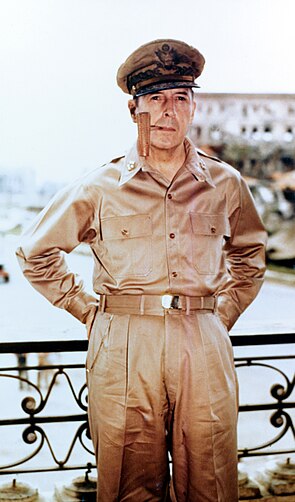 MacArthur in khaki trousers and open necked shirt with five-star-rank badges on the collar. He is wearing his field marshal's cap and smoking a corncob pipe.