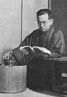 A middle-aged Japanese man, wearing a kimono and glasses, in a kneeling position, is reading a book he holds in his left hand, his elbow resting on a table, while his right hand rests upon a tea kettle
