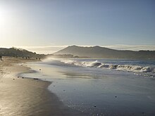 Colour photo of a long beach with foamy waves and mountains in the distance, in the morning light.