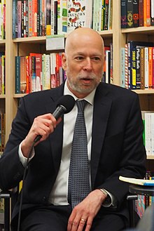 Adam Liptak in a suit and tie, holding a microphone