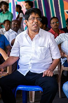 R. Sundarrajan wearing dark pants and a white shirt, sitting on a chair with a group of people, grimacing, looking left of camera