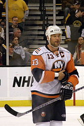 Кавказский мужчина в оранжевой, сине-белой майке. Герен стоит на хоккейной площадке, держит клюшку и прижимает правую руку к животу.