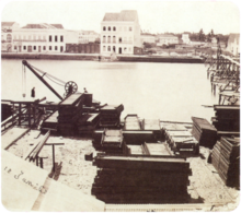 An old photograph showing piles of construction materials and equipment along the bank of a river with large white buildings lining the opposite bank
