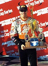 A man in his late twenties holding a trophy in both his hands. He is wearing a white, orange and black racing overalls with sponsors logos and black sunglasses and a black baseball cap.