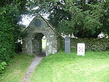 The gate to the churchyard. It is made of cobbled stone and surrounded by overgrown foliage and a low stone wall.