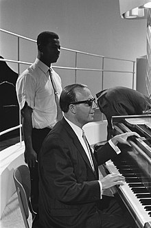 un homme en costume avec des lunettes noires assis au piano, un homme Noir debout derrière lui