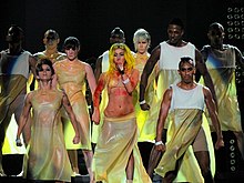 A group of dancers on stage, wearing light yellow colored dresses. In their middle is a blond singer wearing the same dress, with blotches of red color all over her body