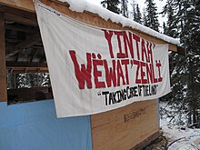 A banner with the words YINTAHʼ WËWATʼZENLÏ in red and beneath, the words "TAKING CARE OF THE LAND" in black